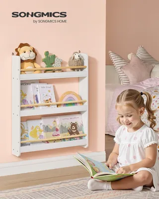 A young girl sits on the floor reading a book next to a white children's bookshelf filled with books and stuffed animals.
