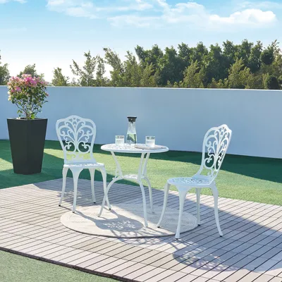 A white aluminum cast bistro set with a table and two chairs on a wooden deck, with a potted plant and a white wall in the background.