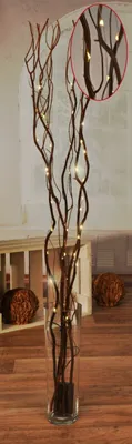 A decorative arrangement of LED-lit branches in a glass vase, placed on a wooden floor with decorative spheres in the background.