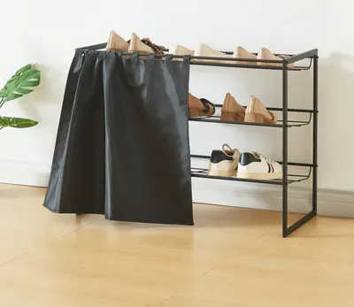 A three-tiered shoe rack with a black fabric cover, holding several pairs of shoes, placed on a light brown wooden floor against a white wall.