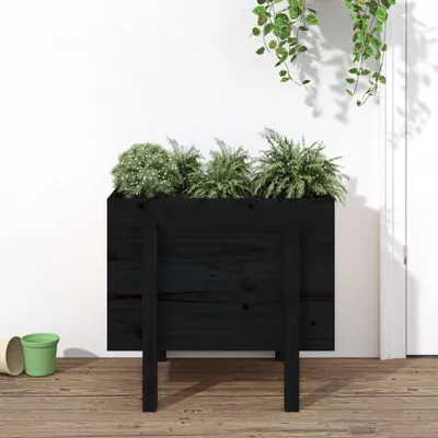 A black wooden planter box filled with green plants, standing on a wooden floor against a white wall.