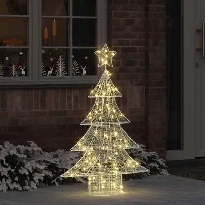 A lighted Christmas tree decoration stands outside a brick building next to a window.