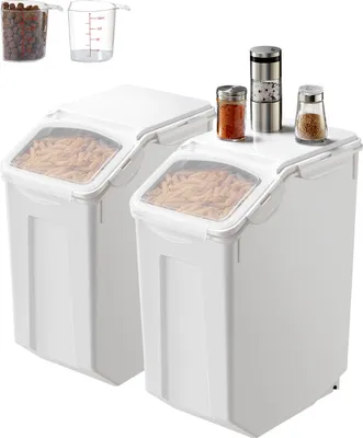 Two white food storage containers with clear lids, filled with pasta, are shown. Small spice jars and a pepper grinder are on top of one container. In the background, two clear measuring cups are visible, one filled with small brown items.