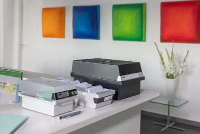 Two card file boxes are on a desk in an office setting, with colorful artwork on the wall behind them.