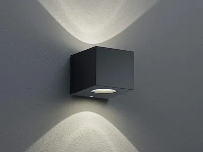 A black cube-shaped wall light mounted on a textured gray wall, casting upward and downward light patterns.