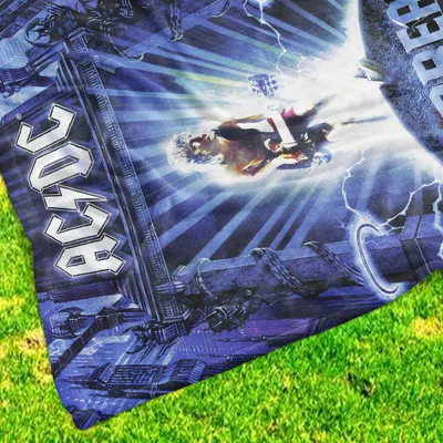 A close-up of an AC/DC branded blanket with a graphic design, laid out on green grass.