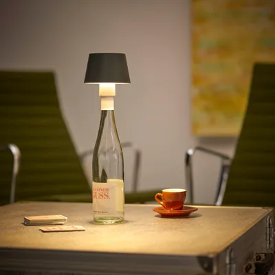 A table lamp made from a wine bottle sits on a table next to a small coffee cup and some cards. Two green chairs are visible in the background.