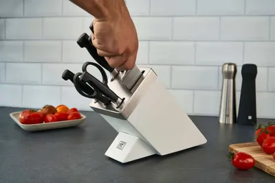 A hand is placing a knife into a white knife block on a kitchen counter, with tomatoes in a dish and salt and pepper grinders in the background.