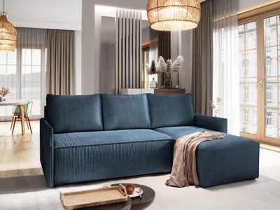 A modern living room with a blue sectional sofa as the main focus, featuring a throw blanket and an ottoman. The room has large windows, curtains, and a woven pendant light.