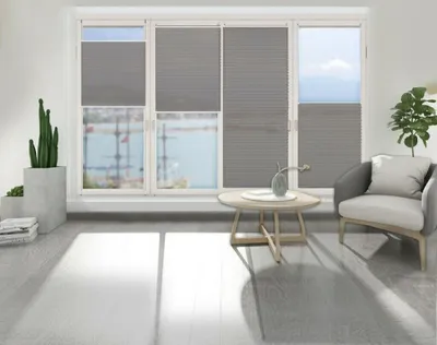 A living room scene with a large window covered by gray pleated blinds, a small round coffee table with a vase, and a comfortable armchair.