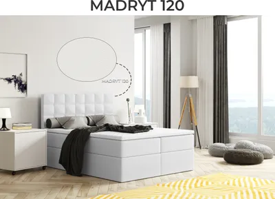 A bedroom scene featuring a white boxspring bed with storage, a headboard, pillows, and a blanket. The room has large windows with curtains, a floor lamp, and a rug.