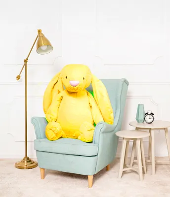 A large yellow plush bunny sits in a light blue armchair next to a gold floor lamp and two small side tables. One table has a vase and an alarm clock on it.