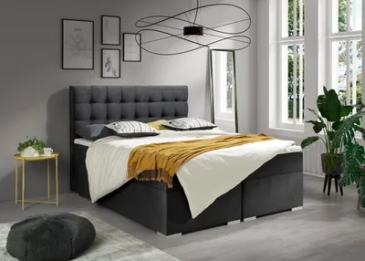A bedroom scene featuring a large bed with bedding and decorative items, set against a grey wall and large windows.