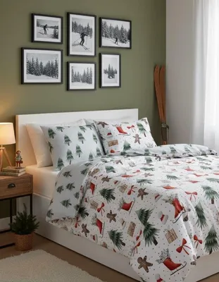 A bedroom scene with a bed dressed in Christmas-themed bedding, framed pictures of skiers on the wall, and a bedside table with a lamp.