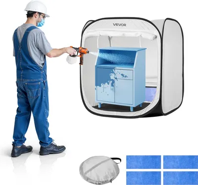 A person in overalls and a hard hat is spraying a blue cabinet inside a pop-up spray booth. The booth has the VEVOR logo on it. Below the booth are a folded carrying case and four blue fabric sheets.