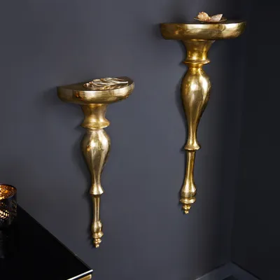 Two ornate gold wall consoles are mounted on a dark gray wall, with a black table and a candle holder in the foreground.