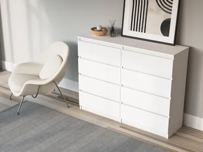 A white dresser with eight drawers sits next to a modern white armchair in a room with light gray walls and a gray striped rug.