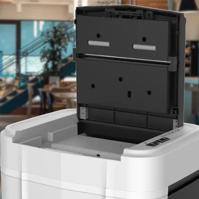 A close-up view of the top of a white and black paper shredder with its lid open, revealing the internal mechanism. The background is blurred and shows a modern interior with stairs and tables, suggesting a commercial or office environment.
