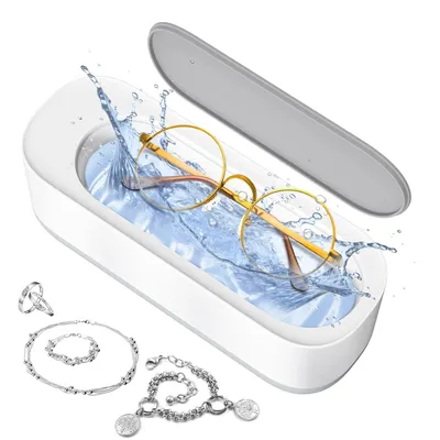 A white ultrasonic cleaner with a gray lid, filled with water and cleaning a pair of gold-rimmed glasses. Several pieces of silver jewelry are placed around the cleaner.