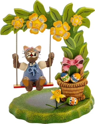 Ein fröhlicher Katzenfigur in blauer Latzhose schaukelt unter einem blühenden Baum während ein Korb mit bunt bemalten Ostereiern und eine gelbe Blume die Szene schmücken