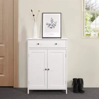 A white cabinet with two doors and a drawer, decorated with a framed picture and a vase with dried flowers, stands in a hallway next to a wooden door and a window. A pair of black boots are placed on the floor next to the cabinet.