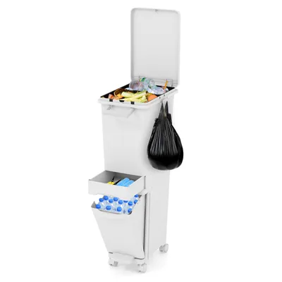 A white, multi-compartment trash can with its lid open, filled with trash. A black trash bag hangs from the side, and a lower drawer contains bottles.
