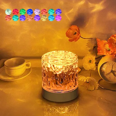 A bedside lamp with a textured glass shade is placed on a surface next to a teacup and saucer, with flowers and a clock in the background. The lamp casts colorful light patterns on the wall.
