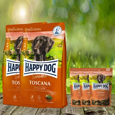 Several bags of Happy Dog dog food are displayed in front of a blurred green background with bokeh lights.