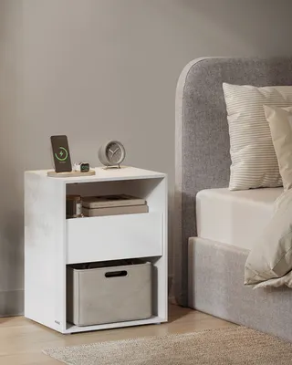 A bedside table with a phone charging, a clock, and books next to a bed.