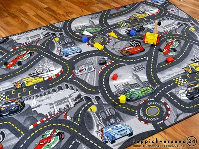 A children's play rug with a race car track design, featuring various toy cars and colorful blocks scattered on it. The rug is placed on a wooden floor.