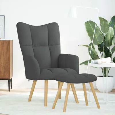 A dark gray fabric armchair with a matching ottoman, placed on a light-colored rug in a well-lit room with a large plant and a white side table.