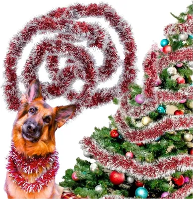 A German Shepherd dog wearing a red and silver tinsel garland around its neck, with a decorated Christmas tree in the background. A large spiral of red and silver tinsel is draped above the dog's head.