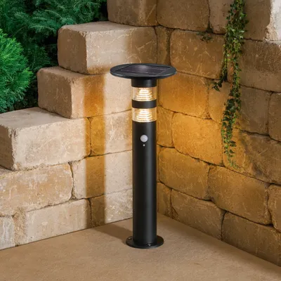 A black solar-powered outdoor light with a motion sensor stands on a patio next to a stone wall and steps.