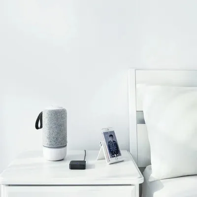 A bedside table with a speaker, a phone, and a small black device.