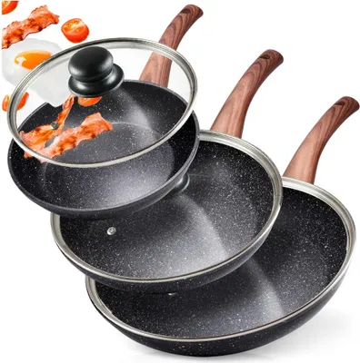A set of three non-stick frying pans with wooden handles and a glass lid, with some bacon and tomatoes in the background.