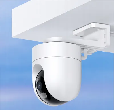 A white security camera is mounted on a white bracket under a white overhang, against a light blue sky.