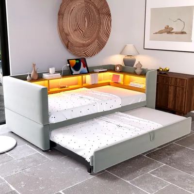 A modern bedroom or living room setting with a light gray upholstered daybed that has a pull-out trundle bed. The daybed features a built-in shelf with LED lighting, a tablet, books, and decorative items.
