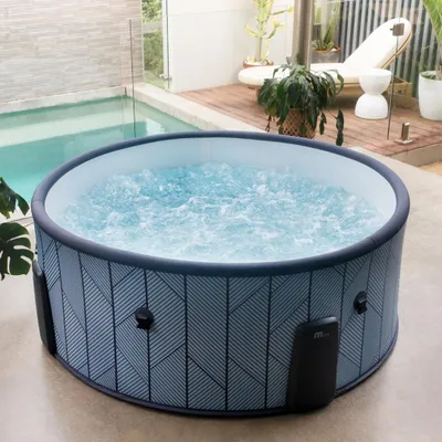An inflatable hot tub filled with bubbling water is placed on a patio next to a swimming pool. The background shows a wooden deck with lounge chairs and plants.