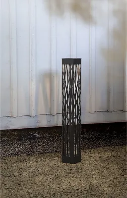 A black solar-powered LED pathway light with a decorative cut-out pattern stands on a gravel surface in front of a white corrugated wall.