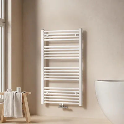 A white towel radiator is mounted on a light beige wall, next to a window and a wooden stool with towels.