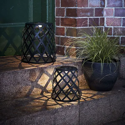 Two black metal lanterns with a diamond pattern are placed on stone steps next to a potted plant. The lanterns are lit, casting a patterned shadow on the steps.