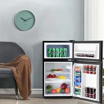 A mini refrigerator with its doors open, showing food and drinks inside, placed next to a chair with a blanket and a clock on the wall.