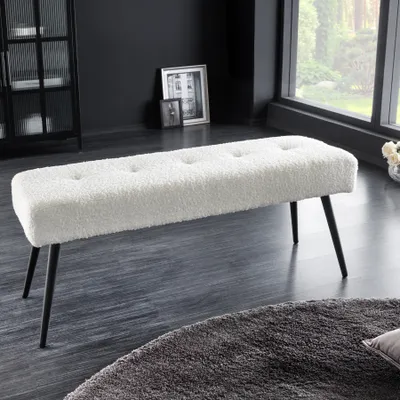 A white bouclé bench with black metal legs is placed on a dark floor in a modern room with a large window and a black cabinet.