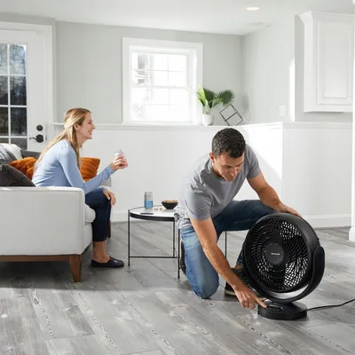 A man is adjusting a fan while a woman sits on a couch holding a drink.