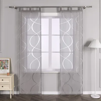 Two sheer grey curtains with a white wavy pattern are hung over a window. A bedside table with drawers and a framed picture is to the left, and a floor lamp is to the right.