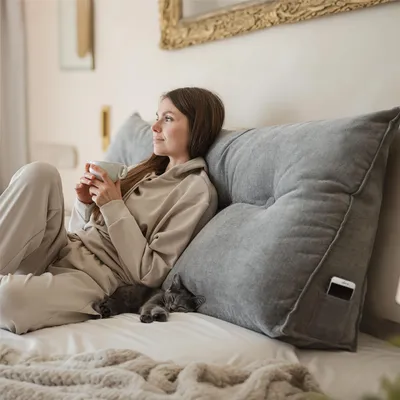 A woman sits on a bed, holding a mug and leaning against a large gray wedge pillow. A small gray cat sleeps on the bed next to her.
