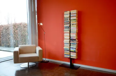 A modern living room with a chair, floor lamp, and a tall, narrow bookshelf filled with books.
