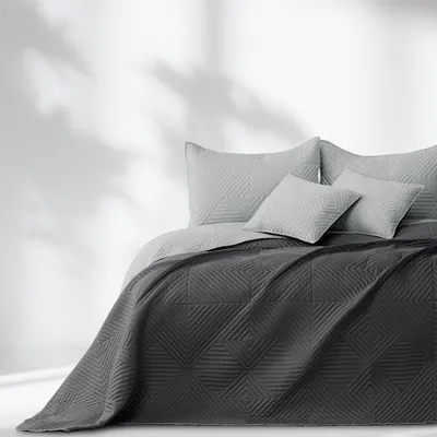 A bed with a dark gray quilted bedspread and light gray pillows, set against a white wall with shadows.