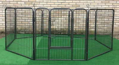An outdoor dog pen is set up on a green surface in front of a brick wall.