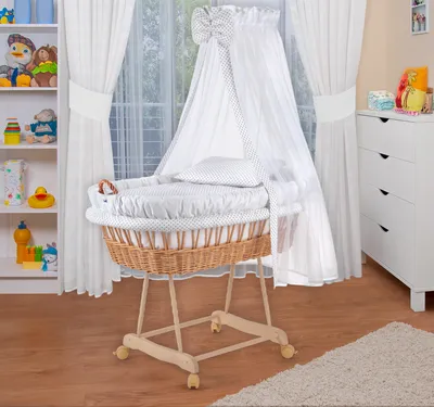 A baby bassinet with bedding and a canopy, set up in a nursery room with shelves of toys and a dresser in the background.